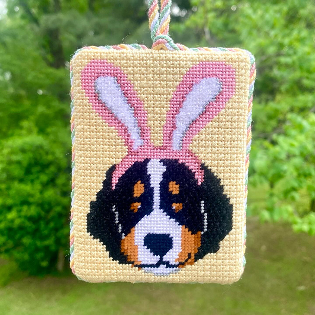 Finished needlepoint ornament of a dog wearing bunny ears, hanging outdoors, from @pinpointneedlepoint on Instagram