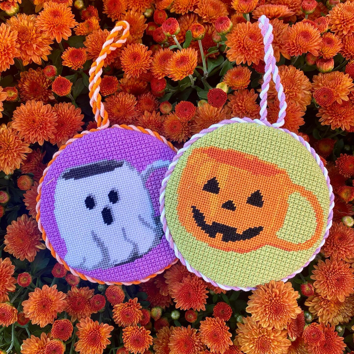 Finished Halloween ghost and pumpkin needlepoint ornaments displayed on orange mums from @pinpointneedlepoint on Instagram